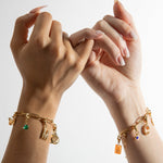 Two hands showcasing gold charm bracelets with various charms including a locket, a heart, and others.