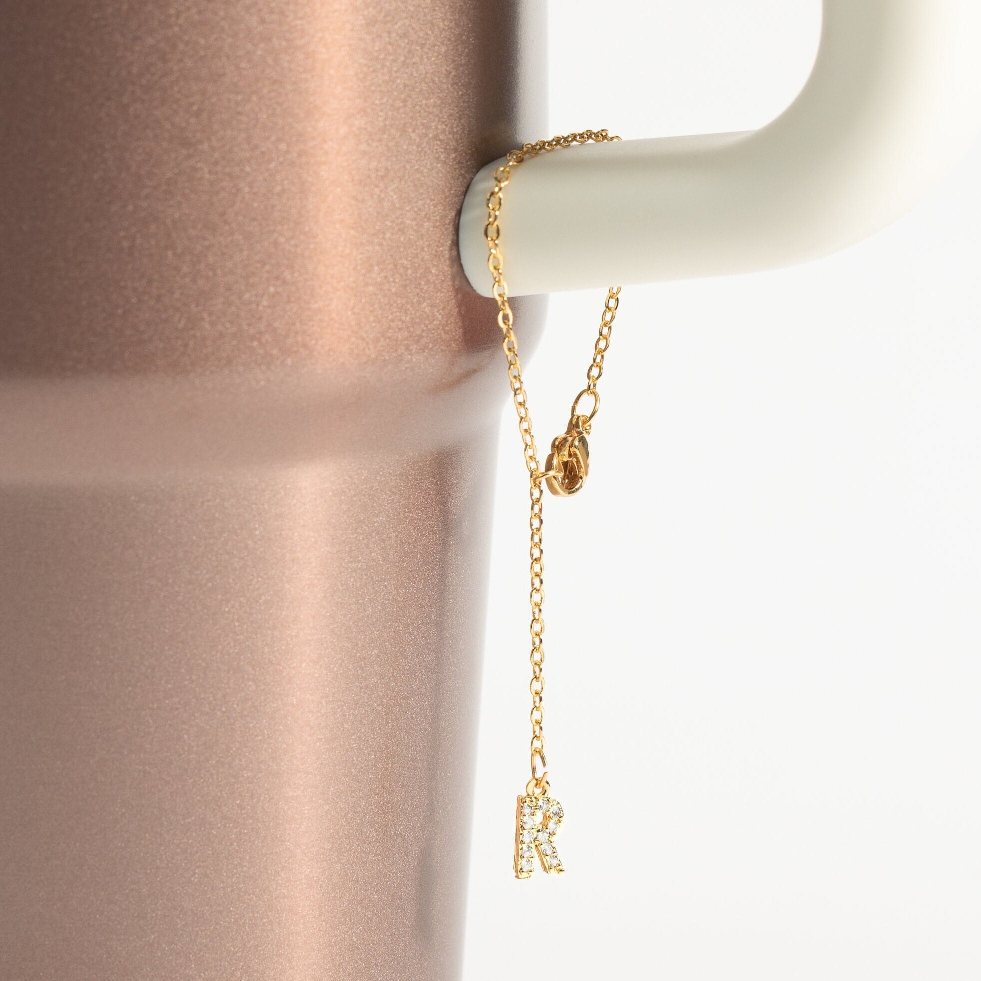 A gold-colored charm in the shape of the Stanley Cup with a personalized initial letter 'R' attached to a white tumbler handle.