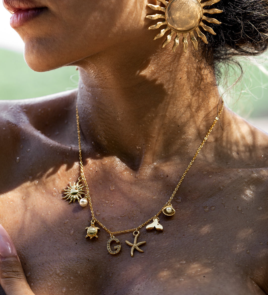 A woman wearing a gold-colored necklace with ocean-inspired charms such as seashells and starfish, and a personalized initial charm.