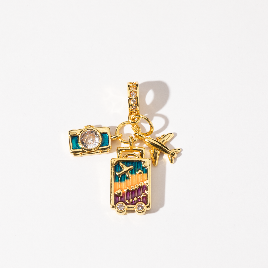 A gold-plated charm featuring detailed charms of a camera, suitcase, and airplane, adorned with vibrant cubic zirconia and sparkling accents.