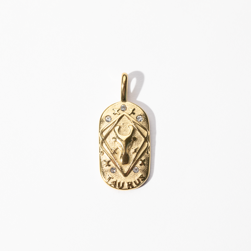A gold-colored engravable Taurus medallion charm with intricate designs and small embellishments.