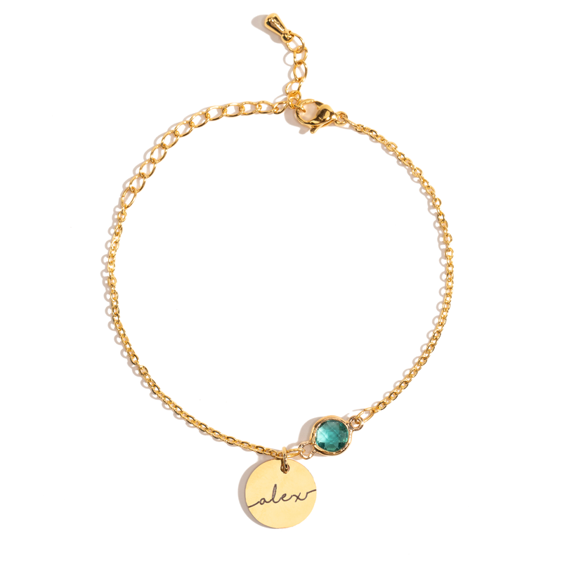 A gold-plated bracelet with a round birthstone charm and an engraved name coin charm on a white background.
