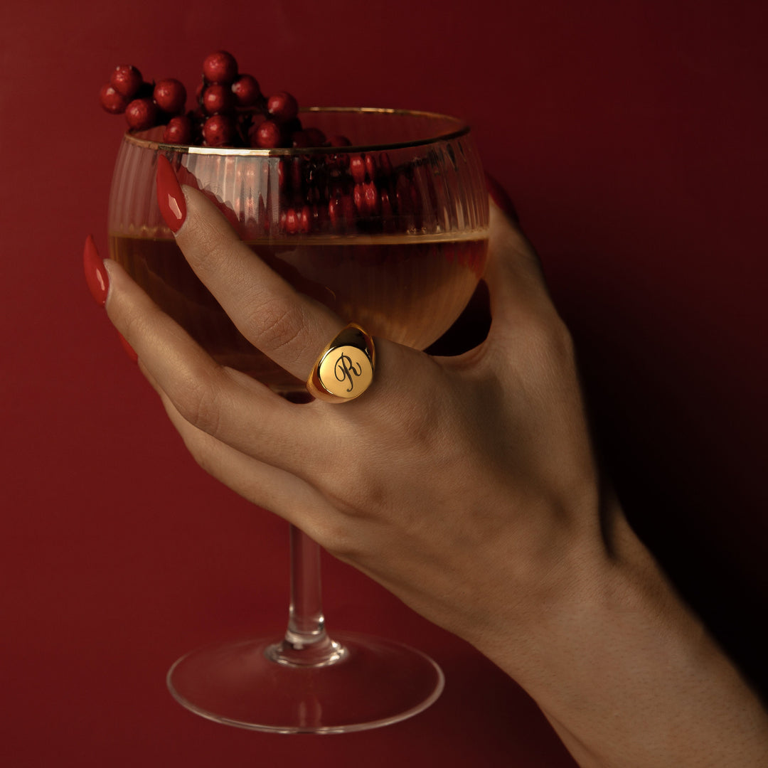 A hand holding a glass of red wine with a custom gold signet ring on the finger, against a red background.