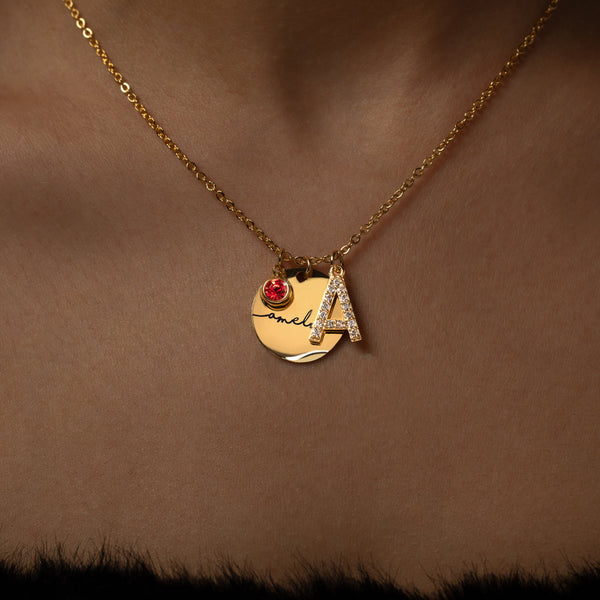 A gold necklace with a personalized initial and birthstone engraving.