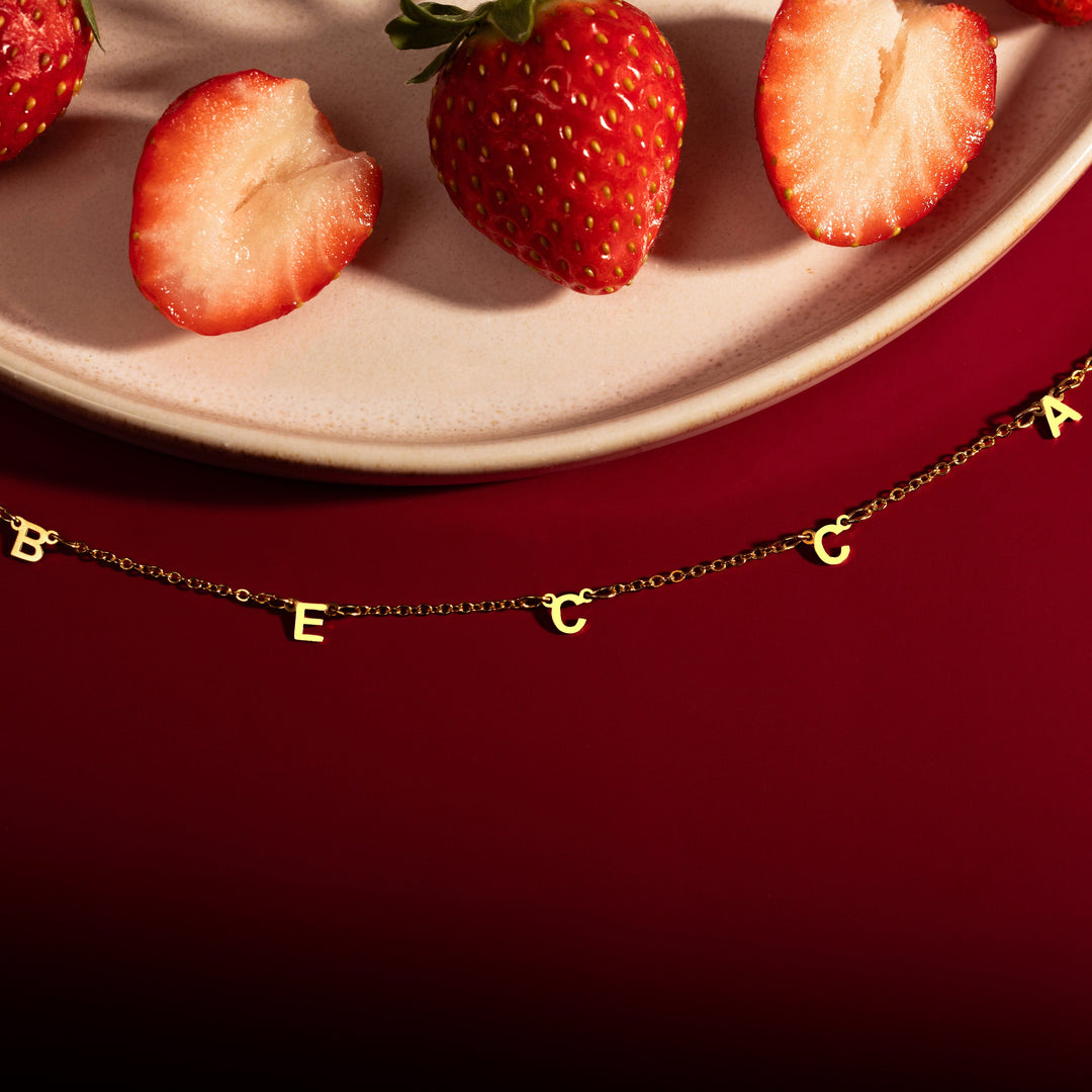 A gold-colored necklace with a personalized message written in block letters, displayed on a red background with strawberries scattered around.