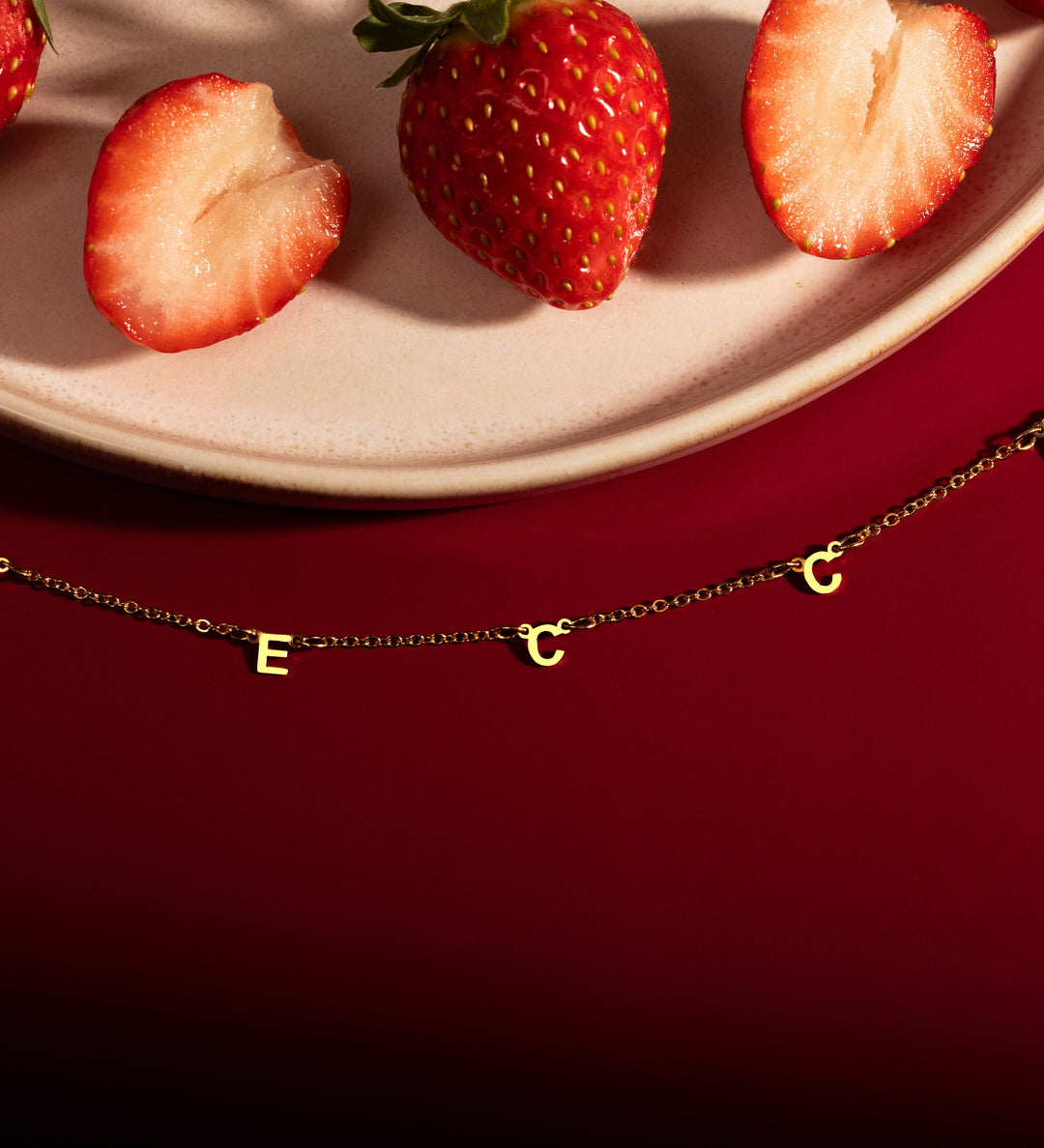 A gold-colored necklace with a personalized message written in block letters, displayed on a red background with strawberries scattered around.