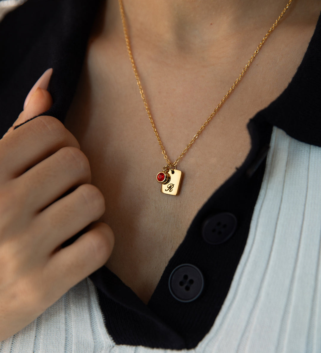 A person wearing a custom gold-colored necklace with an engraved initial 'S' tag attached to a red birthstone, held against a button on a shirt.