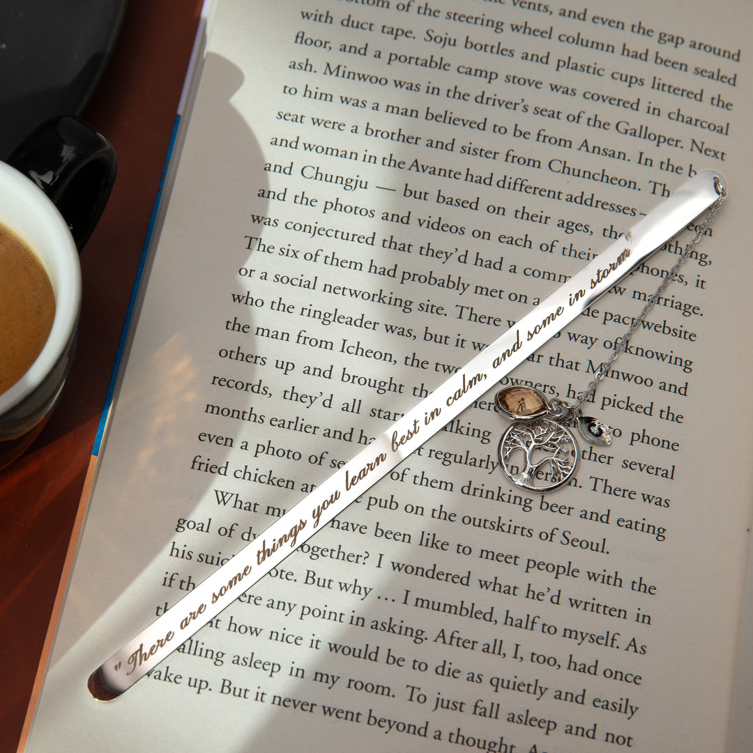 A personalized metal bookmark with a custom birthstone, an engraved initial leaf, and a Tree of Life pendant, displayed on an open book.