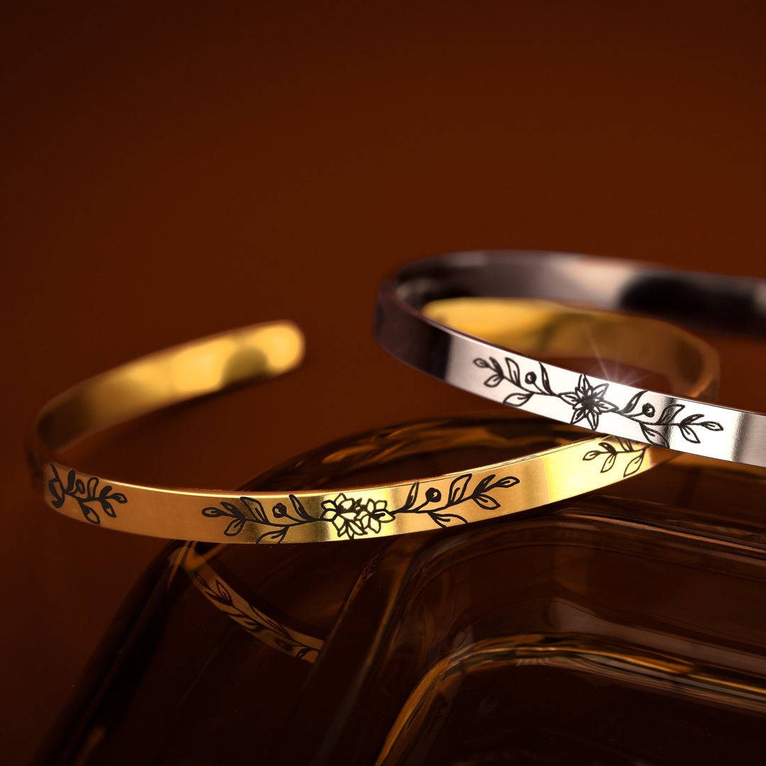 Two bangle bracelets, one in gold and the other in silver, each with an engraved birth flower and leaf design on the front, displayed against an orange-brown backdrop.