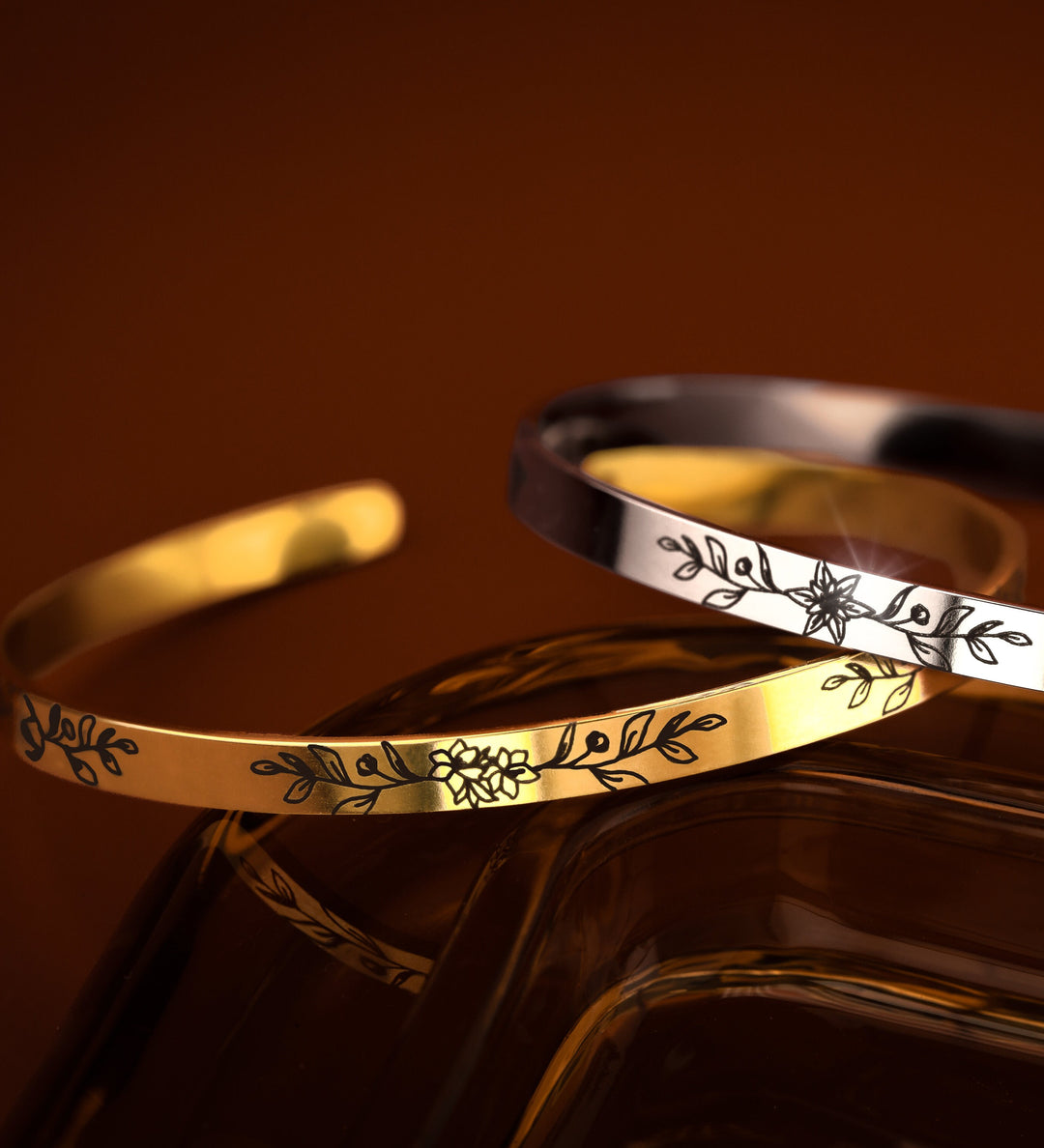 Two bangle bracelets, one in gold and the other in silver, each with an engraved birth flower and leaf design on the front, displayed against an orange-brown backdrop.