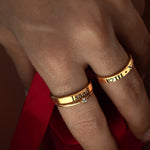 A close-up image of a person's hand wearing two gold-colored rings with engraved text on each.