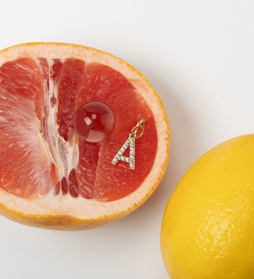 A slice of pink grapefruit with a diamond-encrusted hanging initial letter 'A' piercing earring attached to it, next to a whole lemon.