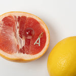 A slice of pink grapefruit with a diamond-encrusted hanging initial letter 'A' piercing earring attached to it, next to a whole lemon.
