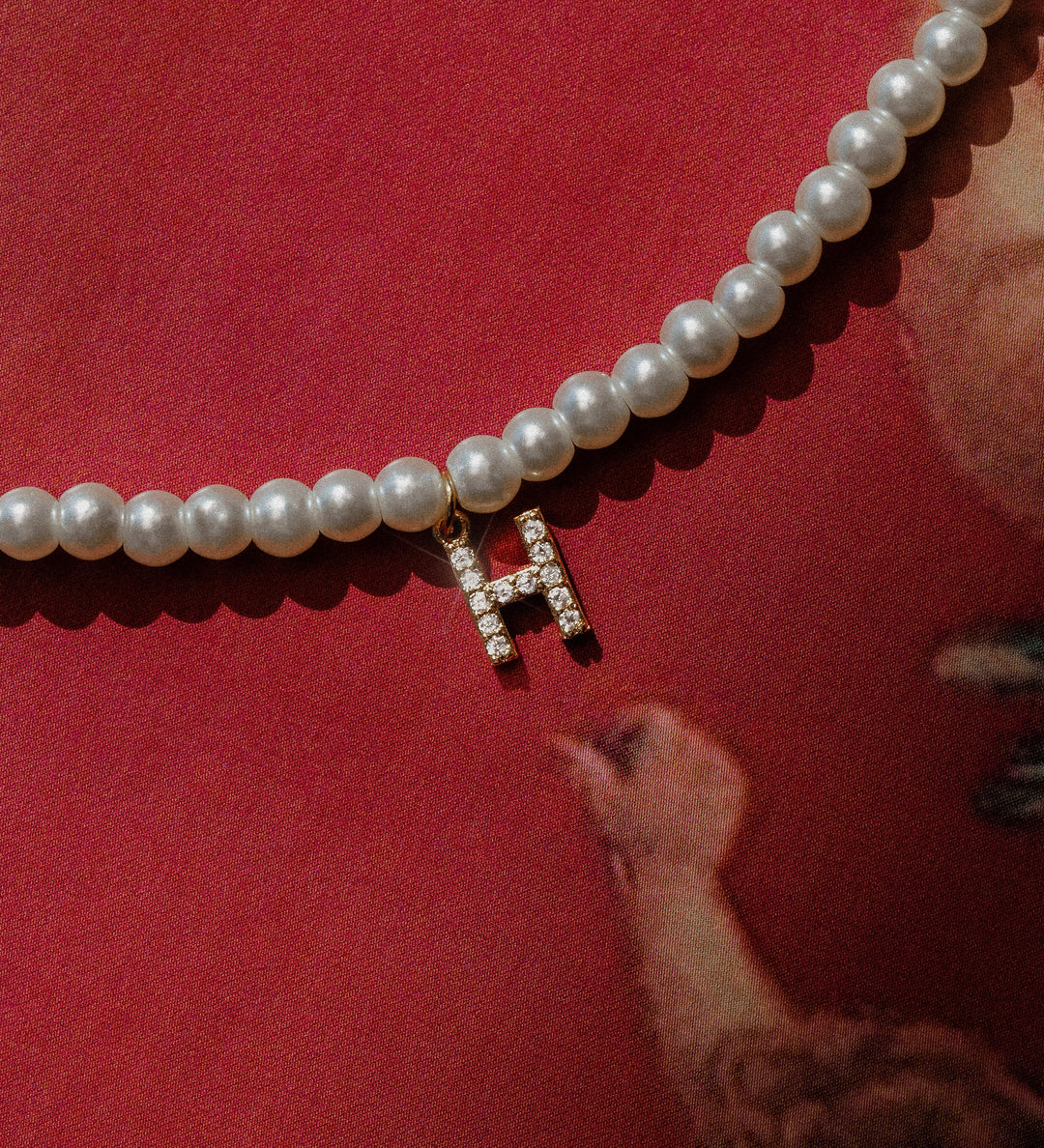 A pearl necklace with a zirconia initial charm resting on a red surface, partially covered by a reflective surface.