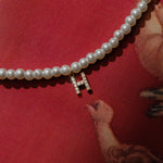 A pearl necklace with a zirconia initial charm resting on a red surface, partially covered by a reflective surface.